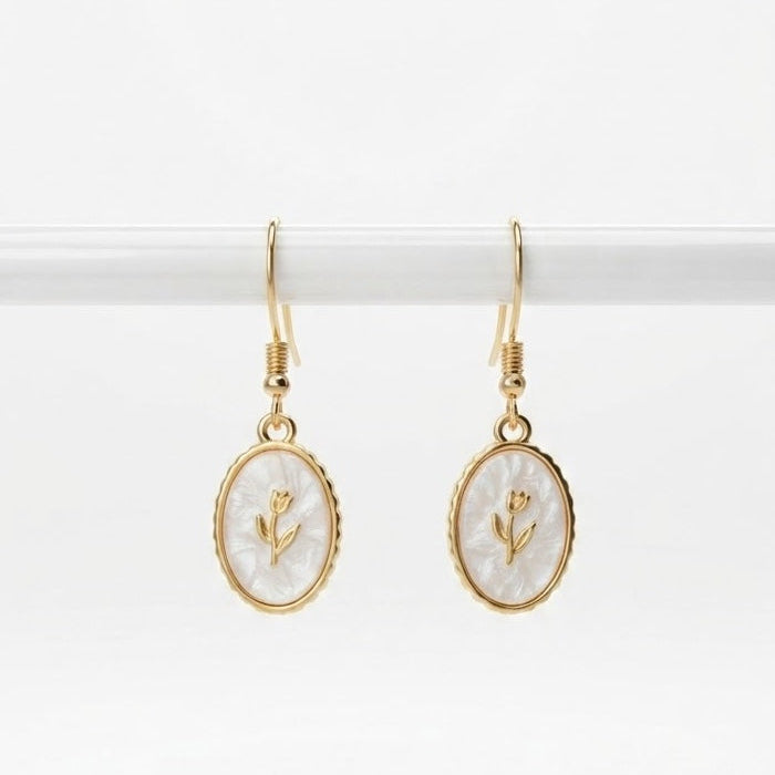 A pair of gold-tone drop earrings with oval white mother-of-pearl style charms featuring a small gold flower design, hanging from gold hooks against a white background.