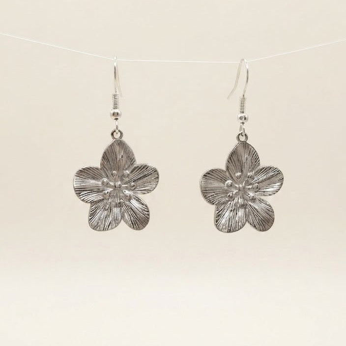 A pair of silver-toned dangle earrings shaped like textured five-petal flowers, hanging from a thin white string against a plain off-white background.