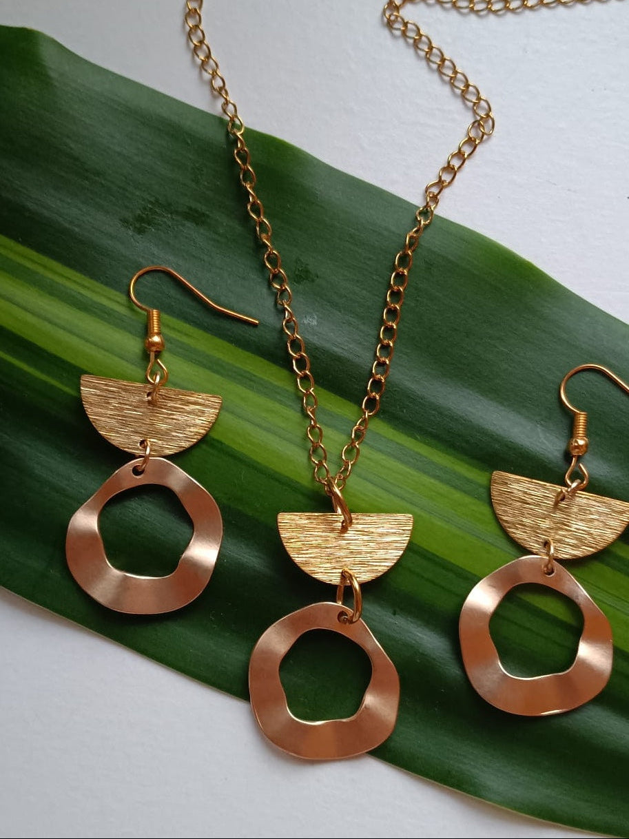 Gold-toned jewelry set with a necklace and two matching drop earrings, styled on a green leaf against a light neutral background.