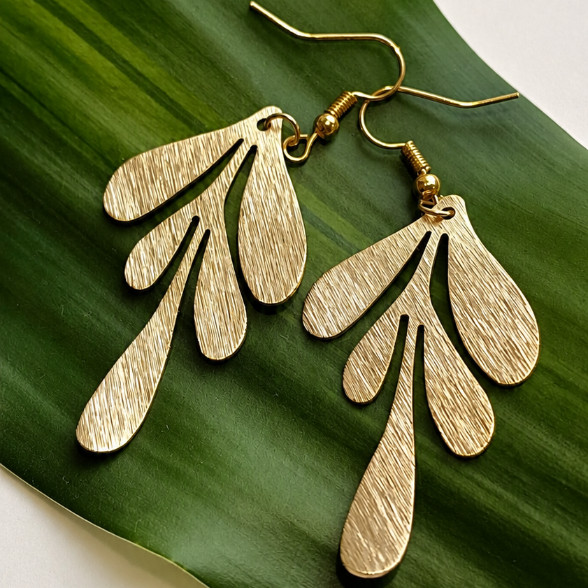 A pair of textured gold dangle earrings with a layered, leaf-like design, displayed on a large green leaf against a light off-white background.
